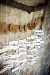 Un ramo di legno da dove penzolano fili con un cuore in legno con su scritto il nome del tavolo e sotto pezzetti di carta con quell degli invitati